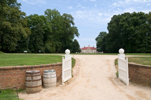 A gravel path leads to a historic mansion, framed by green trees and flanked by two wooden barrels at the entrance.