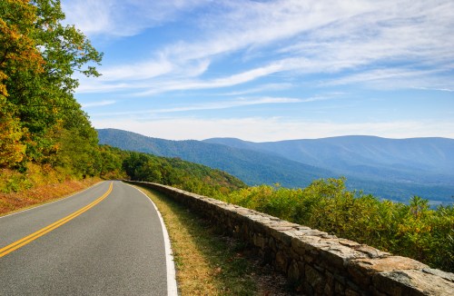 A winding road alongside a stone wall, with mountains and a blue sky in the background, showcasing autumn foliage.