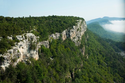 A scenic view of a rocky cliff surrounded by lush green trees and misty mountains in the background.