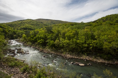 A serene river flows through lush green hills under a cloudy sky, surrounded by trees and rocky banks.