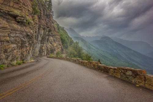 Winding road along a rocky cliff with lush green mountains under a cloudy sky.