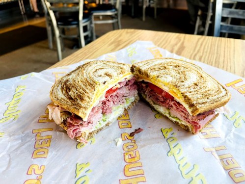 A sliced sandwich with layers of deli meats, lettuce, and tomato on toasted bread, resting on a paper wrapper.