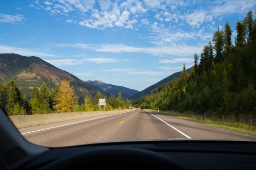 A scenic view of a highway surrounded by mountains and trees under a blue sky with scattered clouds.