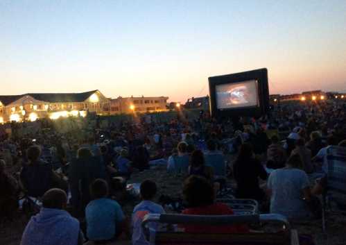 A large crowd sits on the grass watching an outdoor movie on a big screen at dusk.