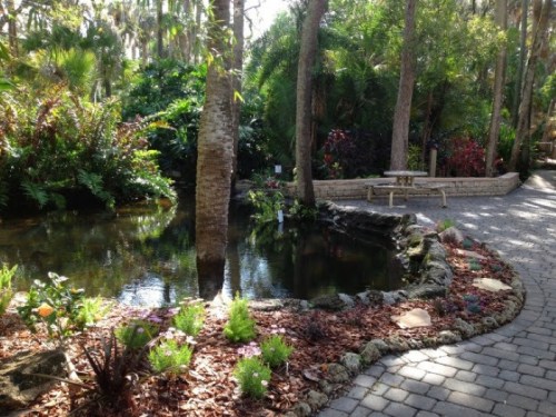 Lush garden with a pond, surrounded by trees and colorful flowers, featuring a stone path and a bench.