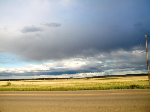 A wide view of a grassy landscape under a cloudy sky, with a road and distant hills in the background.