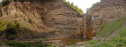 A panoramic view of a waterfall cascading into a serene pool, surrounded by rocky cliffs and lush greenery.