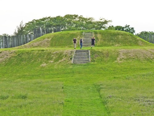Three people walk up grassy mounds with steps, surrounded by greenery and wooden posts in the background.