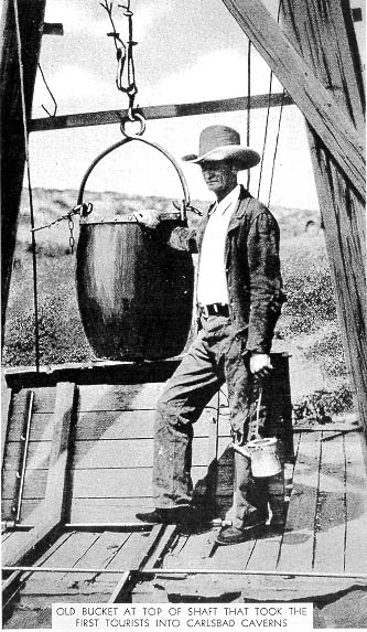 A man stands beside an old bucket at the top of a shaft, used to take the first tourists into Carlsbad Caverns.