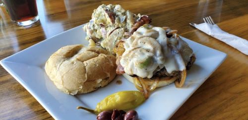 A plate with a burger topped with melted cheese and onions, alongside a serving of potato salad and pickled peppers.
