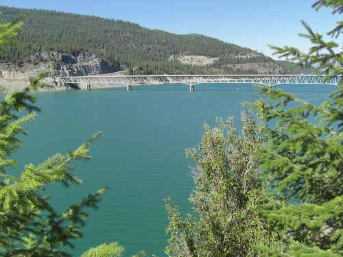 A serene view of a turquoise lake surrounded by green trees and a bridge in the distance, with mountains in the background.