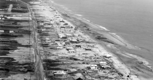 Aerial view of a coastal area showing damaged houses and debris along the beach after a storm or disaster.