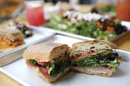 A freshly cut sandwich with layers of greens, tomatoes, and meats, served on a white plate with a colorful salad in the background.