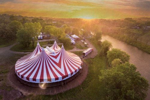 Aerial view of a striped circus tent at sunset, surrounded by trees and a river, with festival setups in the background.