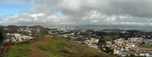 Panoramic view of a city skyline from a hill, surrounded by greenery and cloudy skies.