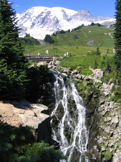 A scenic view of a waterfall cascading down rocks, with lush greenery and a snow-capped mountain in the background.
