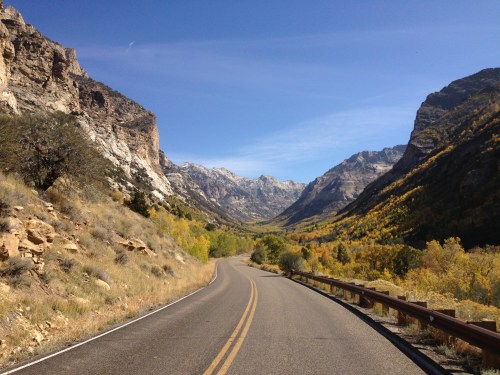 A winding road through a scenic valley surrounded by mountains and autumn-colored trees under a clear blue sky.