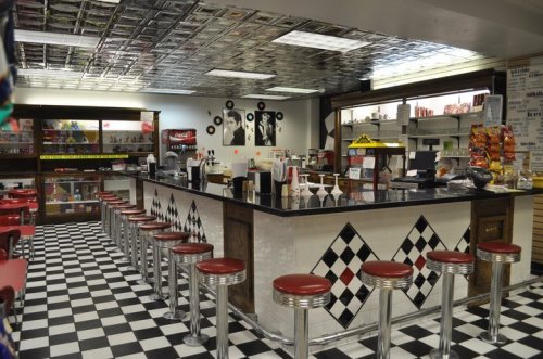 A retro diner interior featuring a checkered floor, a long counter with stools, and vintage decor on the walls.