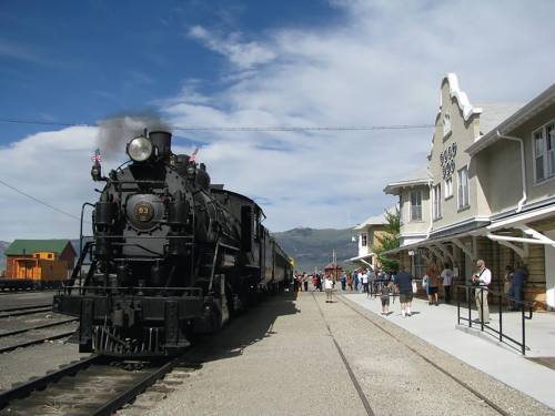 A vintage steam train at a station with people gathered nearby, set against a blue sky and mountains in the background.