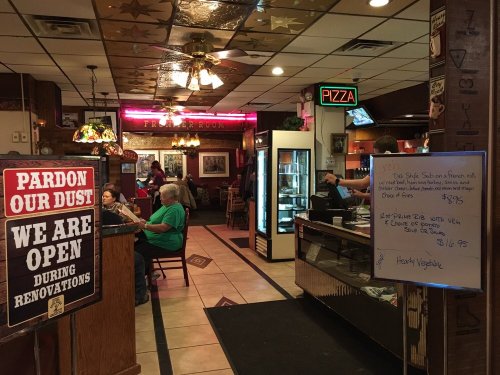 Interior of a restaurant with a "Pardon Our Dust" sign, patrons dining, and a menu board displaying daily specials.