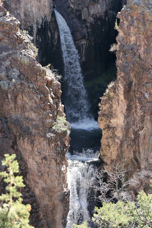 A waterfall cascading between steep rocky cliffs, surrounded by greenery and flowing into a dark pool below.