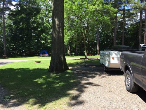 A campsite with a trailer, truck, and a blue tent surrounded by trees and picnic tables on a sunny day.