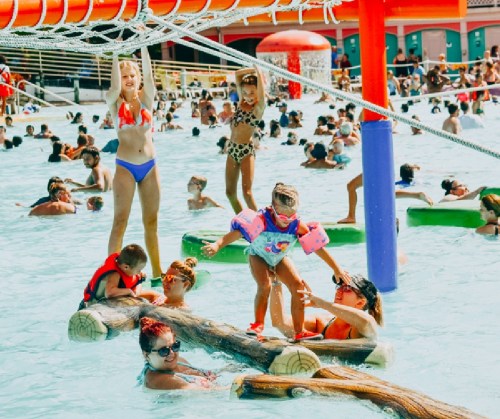 Children and adults enjoy a busy water park, playing on floating logs and in the pool under bright sunshine.