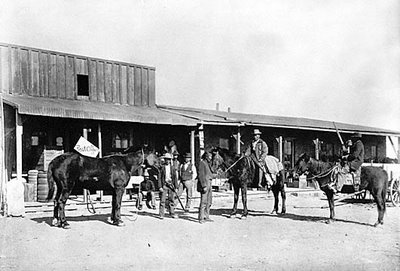 Black and white photo of men on horseback in front of a rustic building, with barrels and equipment nearby.