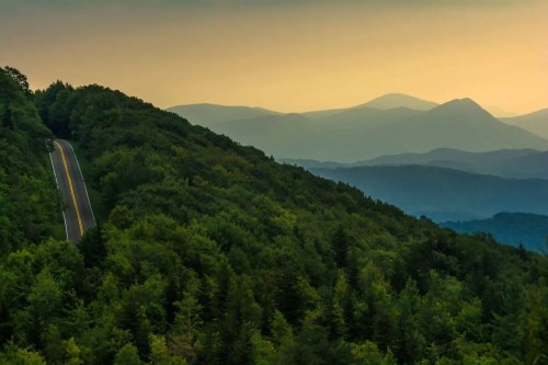 A winding road through lush green hills, with distant mountains under a soft, hazy sunset sky.
