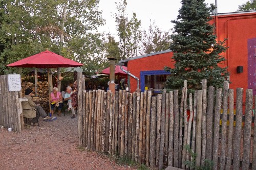 A cozy outdoor scene with people sitting under red umbrellas near a colorful building and a wooden fence.