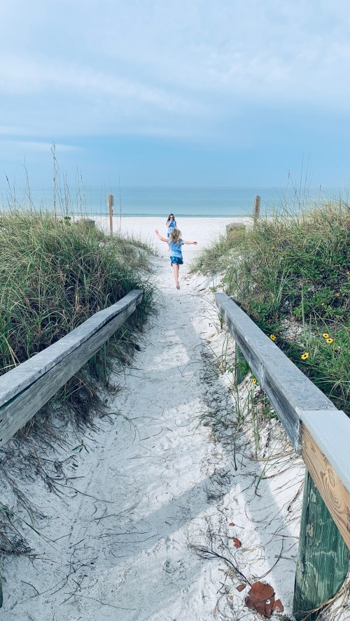 A child runs joyfully down a sandy path towards a beach, surrounded by tall grass and a cloudy sky.