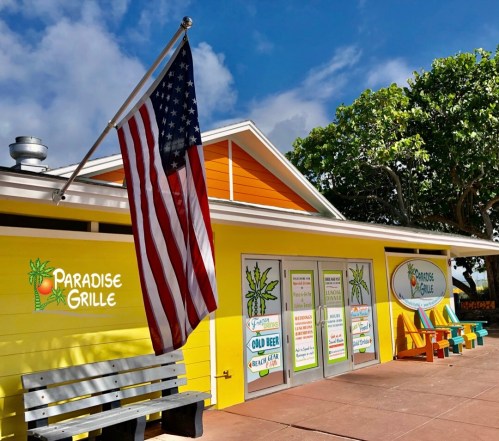 A colorful restaurant exterior with an American flag, featuring signs for food and drinks, surrounded by trees.