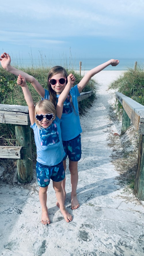 Two girls in matching blue outfits and sunglasses pose joyfully on a sandy path leading to the beach.