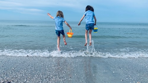Two children jump into the water at the beach, holding colorful buckets, with gentle waves splashing around them.