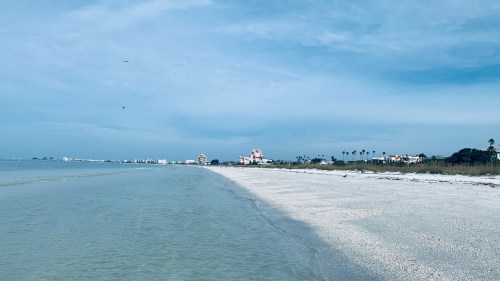 A serene beach scene with gentle waves, soft sand, and distant buildings under a cloudy sky.