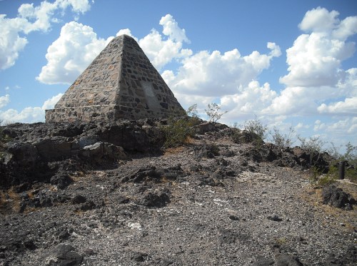 A stone pyramid on a rocky hilltop under a blue sky with fluffy clouds. Sparse vegetation surrounds the area.
