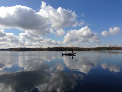 A lone boat on a calm lake reflects fluffy clouds and trees under a blue sky.