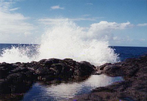 Waves crashing against rocky shore under a blue sky with scattered clouds.