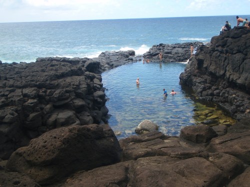 A natural rock pool by the ocean, with people swimming and relaxing among dark volcanic rocks.