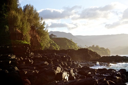 A rocky coastline with lush greenery and a distant house, illuminated by soft sunlight and a cloudy sky.
