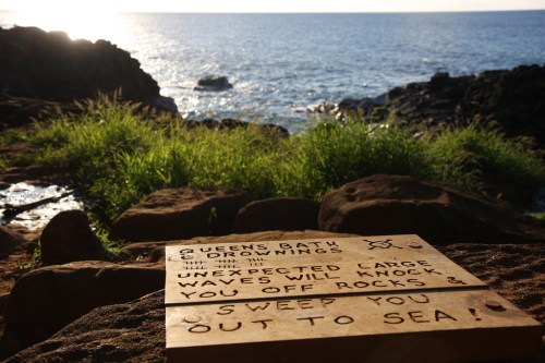 A wooden sign warns of dangers at Queens Bath, with ocean waves crashing against rocks in the background.