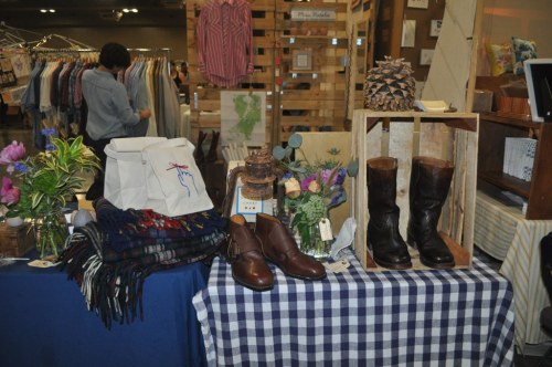 A display of clothing, boots, and accessories on tables, with flowers and a wooden crate in a market setting.