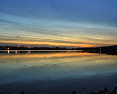 A serene lake at sunset, reflecting colorful skies and soft clouds, with gentle ripples on the water's surface.