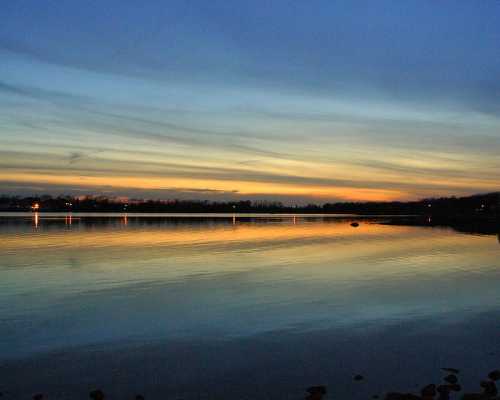 A serene lake at sunset, reflecting colorful skies and soft clouds, with gentle ripples on the water's surface.