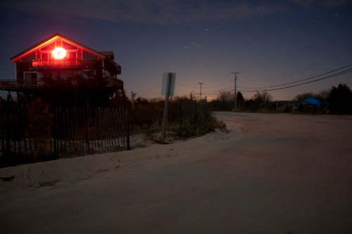 A house with a red light in the window stands by a quiet road at night, surrounded by sparse vegetation and stars.
