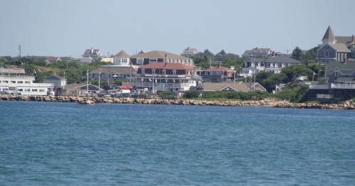 Coastal view featuring houses along the shore, with rocky outcrops and clear blue water under a sunny sky.
