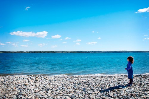 A child stands on a rocky beach, gazing at the calm blue water under a clear sky with a few clouds.