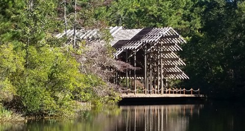 A wooden structure with a slatted roof beside a calm, reflective pond, surrounded by lush greenery.