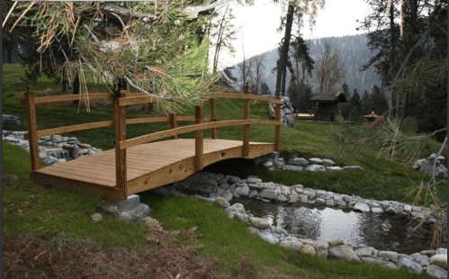A wooden bridge over a small stream, surrounded by greenery and trees in a serene outdoor setting.