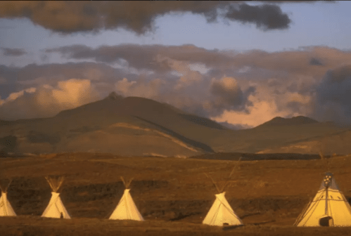 A serene landscape featuring several white tipis against a backdrop of mountains and a colorful sky at sunset.